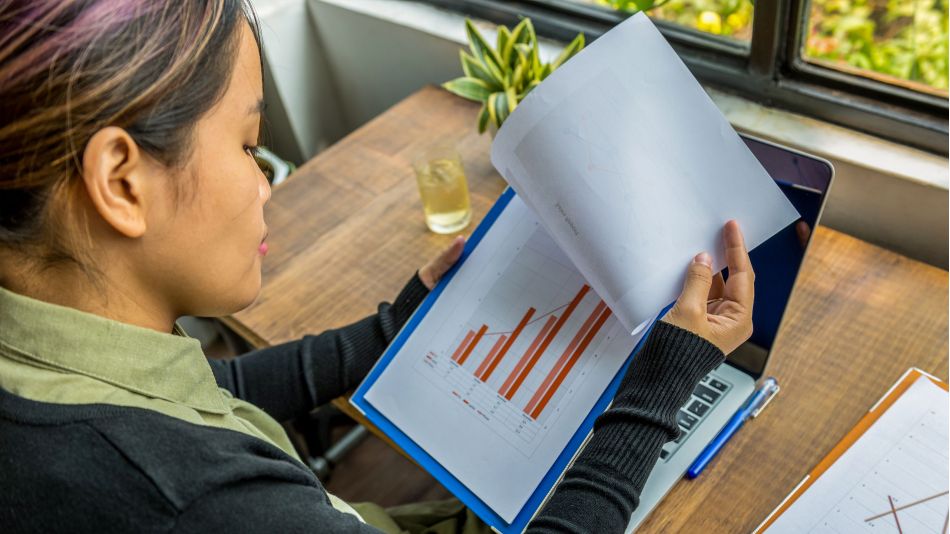 Woman reviewing property report documents and charts at a desk beside a laptop and window